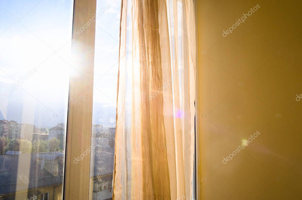 Sun shining through curtains and window. Sunlit apartment on sunset. City view and blue sky.