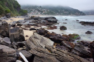 Okyanus sahilindeki dev kayalar. Sisli bulutlu hava. Arka plandaki tepede küçük bir köy. Maicolpue, Osorno, Pasifik Sahili, Şili.