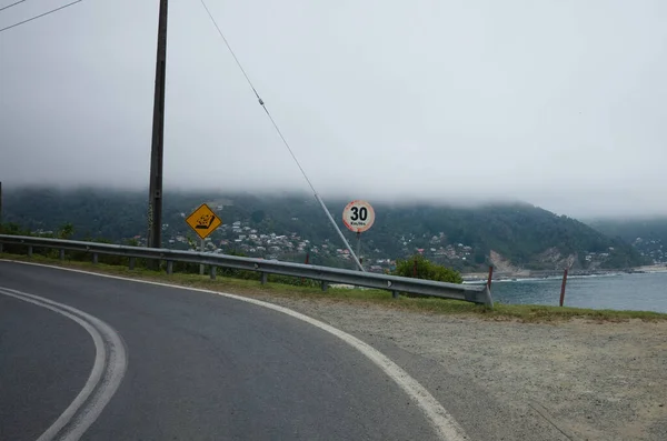 Sharp turn road before cliff and speed limit sign behind. Warning sign falling rocks. Ocean beach and village on background. Chile