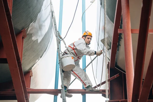 Industrial climber in helmet and uniform painting water tower. Professional Painter working on height. Risky job. Extreme occupation.