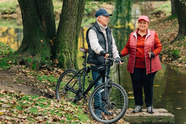 Pretty senior sport couple standing  in colorful autumn park near river. Mature woman with nordic walking poles and old man with bicycle resting outdoors.