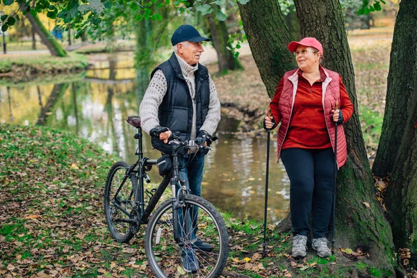 Pretty senior sport couple standing  in colorful autumn park near river. Mature woman with nordic walking poles and old man with bicycle resting outdoors.