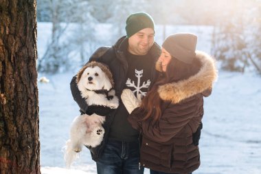 Sevimli Genç çift ile onların küçük beyaz köpek kış ormandaki eğleniyor. Erkek ve kadın kazaklı bir kar tanesi ile
