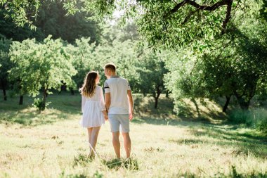 Parkta yürüyen aşık genç güzel çiftin arka görünümü. Onun erkek arkadaşı sarılma beyaz elbiseli yakışıklı neşeli sarışın kız. Dışarıda eğlenen kadın ve erkek
