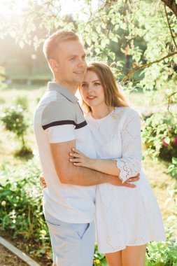 Genç çift aşk ayakta parkta güzel. Beyaz elbiseli erkek arkadaşıyla sarılma yakışıklı neşeli sarışın kız. Erkek ve kadın dışarıda eğleniyor