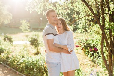 Genç çift aşk ayakta parkta güzel. Beyaz elbiseli erkek arkadaşıyla sarılma yakışıklı neşeli sarışın kız. Erkek ve kadın dışarıda eğleniyor