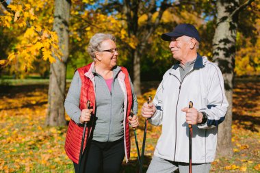 Renkli sonbahar parkında İskandinav yürüyüş direkleri olan oldukça yaşlı bir çift. Olgun kadın ve yaşlı sporcu dışarıda dinleniyor.. 