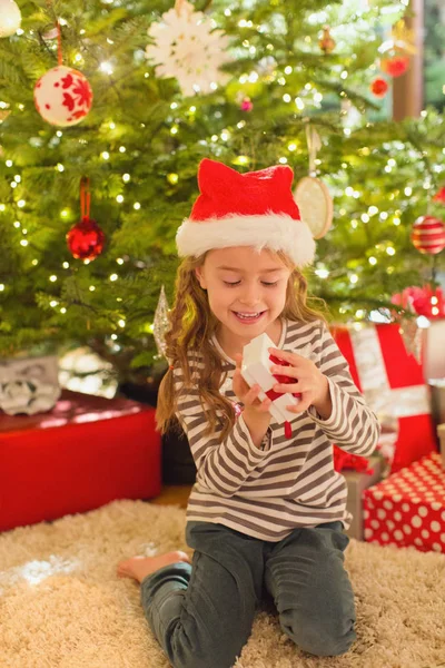 Lächelndes Mädchen Mit Weihnachtsmütze Öffnet Geschenk Vor Dem Weihnachtsbaum — Stockfoto