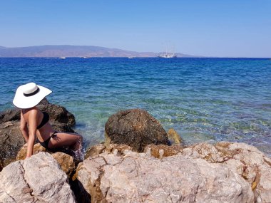 Bir kadın beyaz hasır şapkalı taşlı deniz kıyısında oturan ve gemi geçen deniz horizon adlı arıyorsunuz