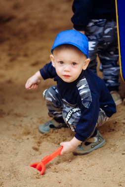 Bir çocuk oyun alanında kumla oynuyor ve elinde bir kürek tutuyor.