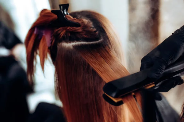 Peluquería alisa el cabello pelirrojo de la mujer con herramienta de alisador de pelo en el salón de belleza . — Foto de Stock