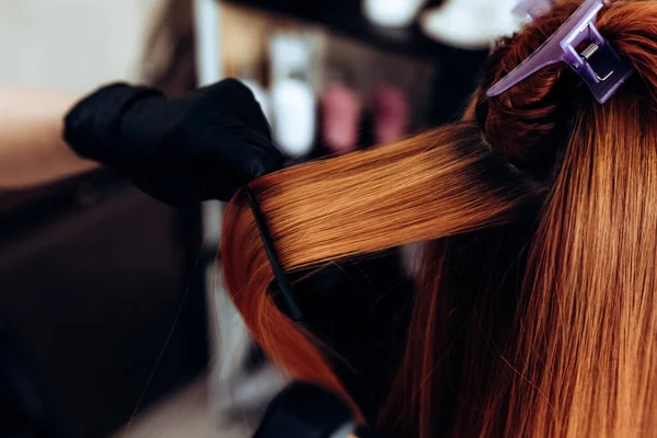 Fryzjer prostuje rude włosy kobiety z prostownicą do włosów w salonie fryzjerskim. — Zdjęcie stockowe