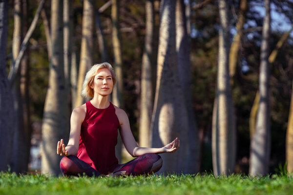 Beautiful people Fitness woman with short blonde hair doing yoga,concentrate and smile in a park of bottle trees, sitting on the grass. Sport and Healthy Lifestyle Concept