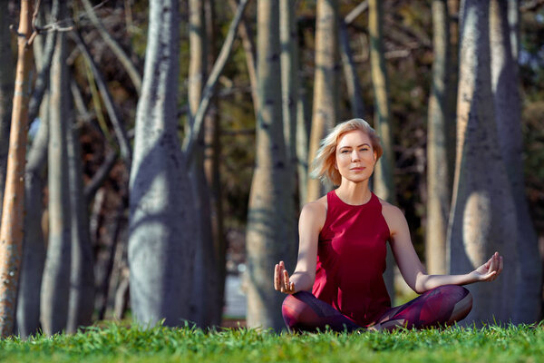 Beautiful people Fitness woman with short blonde hair doing yoga,concentrate and smile in a park of bottle trees, sitting on the grass. Sport and Healthy Lifestyle Concept