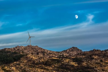View of a wind turbine on top of mountains, dramatic sunset sky with a moon as background, in Portugal...
