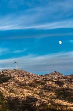View of a wind turbine on top of mountains, dramatic sunset sky with a moon as background, in Portugal...