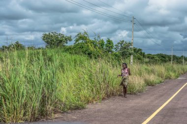 Malange / Angola - 12 08 2018: Malange / Angola - 12 08 2018: bitki örtüsü ve arka plan olarak bulutlu gökyüzü ile yol boyunca yürüyüş bir kız görünümünü