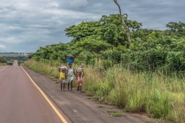 Malange / Angola - 12 08 2018: Çocuklu kadınla görünümünü arka plan olarak tropikal peyzaj boyunca yol kenarında kafa yük ile yürüyor