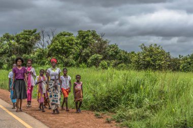 Malange / Angola - 12 08 2018: View of a group at young girls walking along roadside, tropical landscape as background clipart