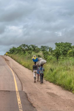 Malange / Angola - 12 08 2018: çocuklar görünümünü arka plan olarak tropikal peyzaj boyunca yol kenarında kafa yük ile yürüyor