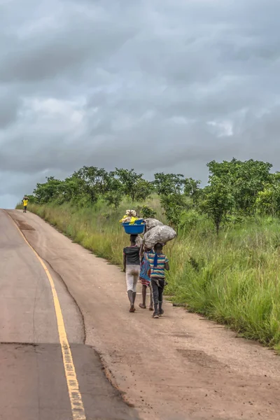 Malange / Angola - 12 08 2018: çocuklar görünümünü arka plan olarak tropikal peyzaj boyunca yol kenarında kafa yük ile yürüyor