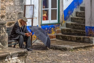 Ortaçağ köyü Obidos'un meydanında, yerde yağmur şapkası açık, ahşap bankta oturan, tipik paltoile yaşlıgörünümü