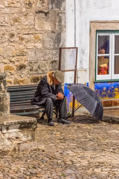 View of elderly with typical coat, sitting on wooden bench, with rain hat open on the ground, in the square of the medieval village of Obidos Stock Image