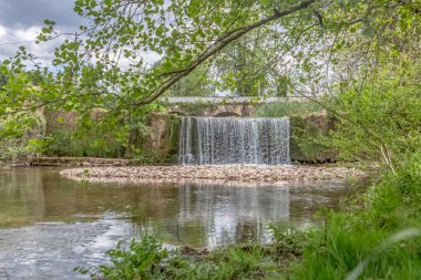 Kıyılarda ağaçlar, kayalar ve bitki örtüsü ile nehir üzerinde küçük bir çağlayan görünümü