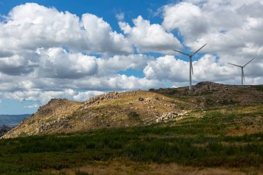 Dağların tepesindeki rüzgar türbinlerinin manzarası, toprak yol ve vahşi doğa ve manzara, arka plan olarak bulutlu gökyüzü...