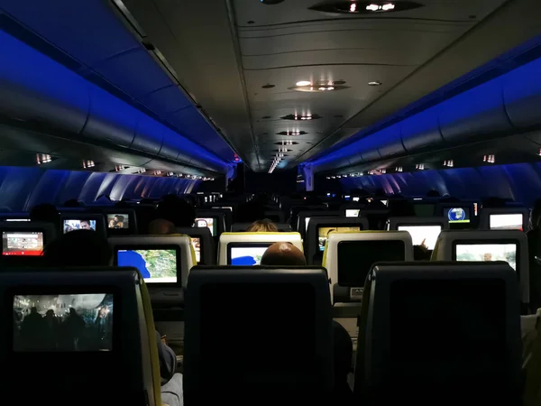 View from inside the plane in mid-flight, low lights, with blue LED and emergency light, scenery with rear perspective of the seats with people watching TV and sleeping...