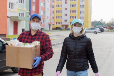 Koruyucu maskeli gönüllüler bir kutu yiyecekle caddede yürürler.