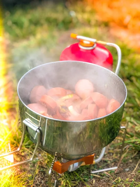 Camping cooking set of a burner, gas bottle and a pot used for preparation of a meal from sausages and an onion outdoors.