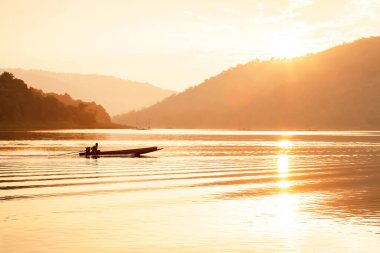 Erkek Asyalı balıkçı, alacakaranlıkta gölde ahşap balıkçı teknesini kullanıyor. Güneş arka planda dağın arkasında batıyor. Gölün yüzeyinde ve dalgalarda parlıyor..