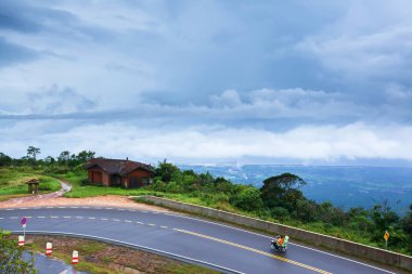 Khmer ailesi yağmurlu bir günde dağ asfalt yolunda motosiklet sürerken arka plandaki körfezi bulutlu kapladı. Kampot, Güney Kamboçya.