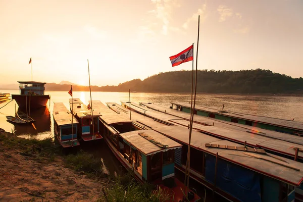 Gün batımında Mekong Nehri 'nin sakin bir manzarası, arka planda bir dağ sırasının ardında parlayan güneş, nehir kenarında bir sürü geleneksel ahşap tekne. Luang Prabang, Kuzey Laos.
