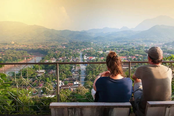 Back view of couple young European travelers are sitting on marble chair while enjoying view Luang Prabang ancient town at sunset from the top of Mount Phou Si, travel destinations in Laos. UNESCO World Heritage Site.