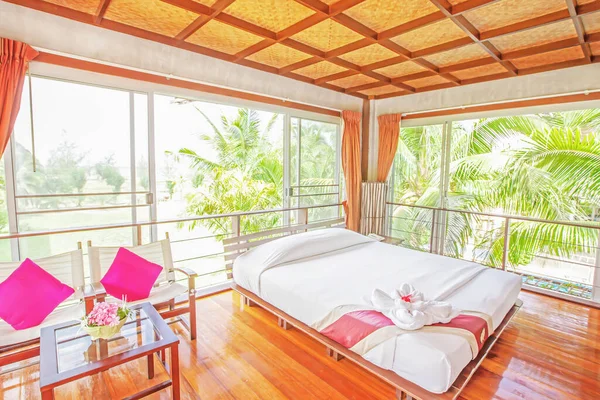 Picturesque interior of bedroom, sunrise shines through broad windows down on glossy wooden floor, palm trees and tropical garden in the courtyard. High-key lighting. Soft focus on the bed.