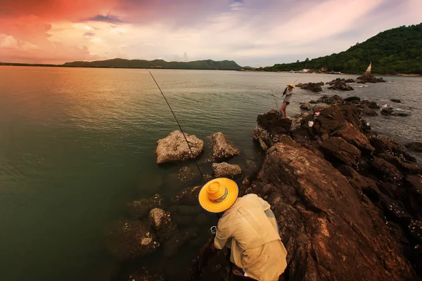 Two Asian fishermen are fishing off the rock on a seashore at dusk, dramatic clouds and sunset sky in the backgrounds. Focus on the fisherman.