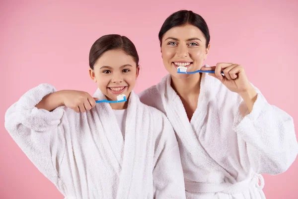 Mother and daughter in gowns brushes teeth.