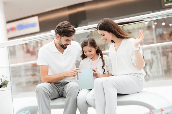Família, pai, mãe e filha estão sentados no banco do shopping. Menina está olhando dentro de um saco . — Fotografia de Stock