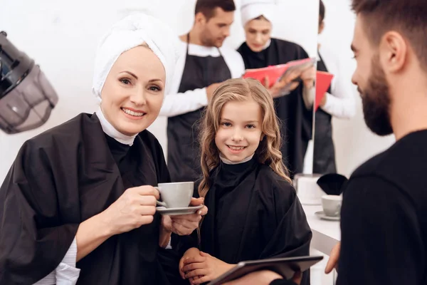 Mulher madura com xícara de café e pequena menina encaracolada fazer penteados no salão de cabeleireiro . — Fotografia de Stock