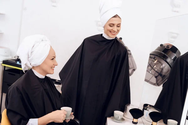Two adult women with towels on their heads drinking coffee at hairdresser\'s salon.