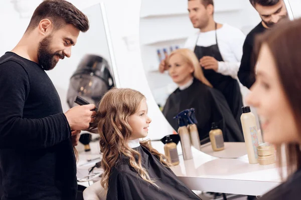 Smiling hairstylist combs curly hair of cute little girl in beauty salon. Cute little girl doing stylish haircut in beauty salon.