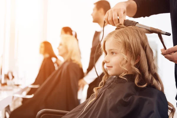 Barbero barbudo hace rizos en el pelo de hermosa niña con rizador de pelo . —  Fotos de Stock