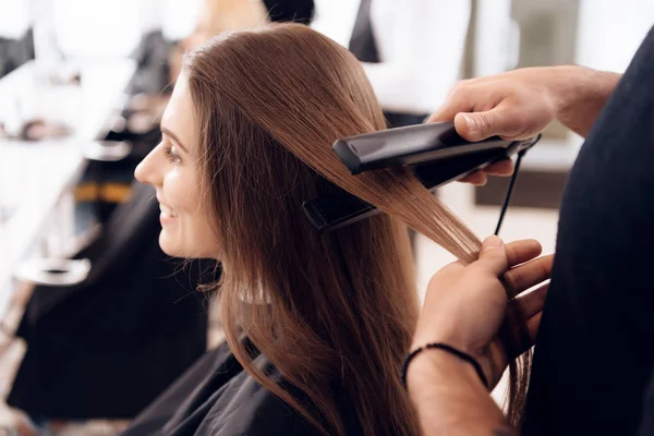 Female hairdresser straighten brown hair of beautiful woman using hair tong in beauty salon.