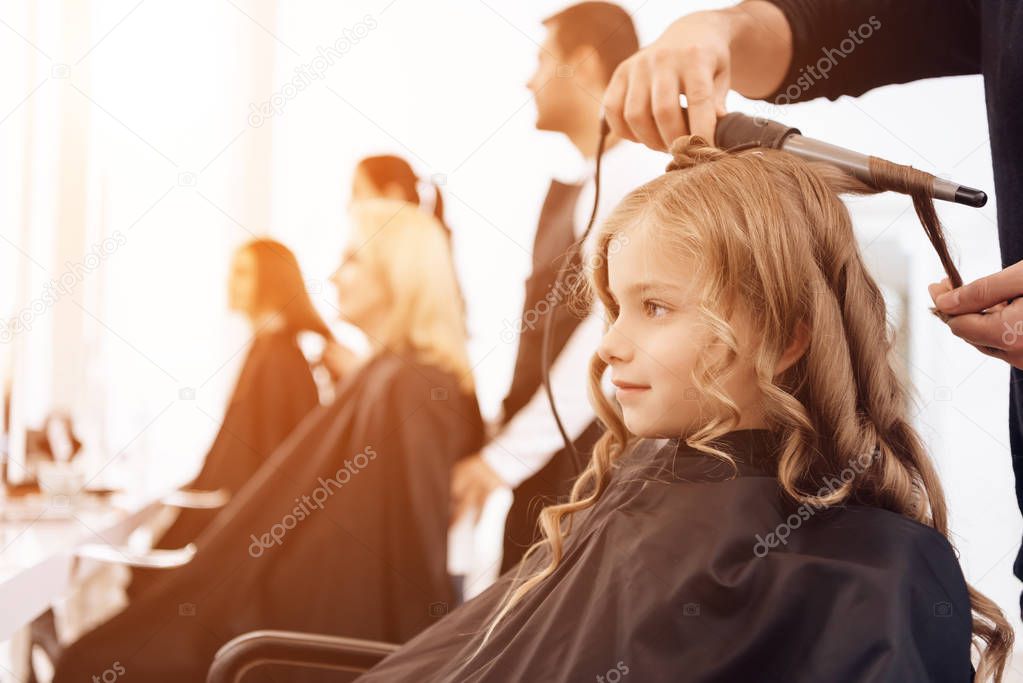 Bearded barber makes curls on hair of beautiful little girl with hair curler.