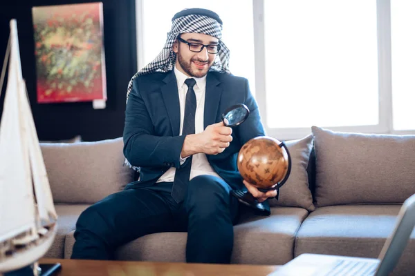 Arab businessman looking at globe with loupe on couch at hotel room. — Zdjęcie stockowe