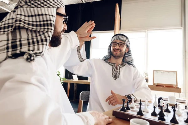 Two arab businessmen high five behind chessboard at hotel room. — Stockfoto