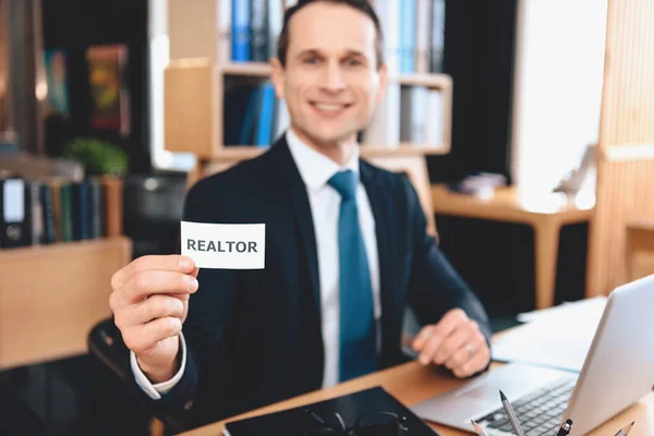 Agente inmobiliario sentado en el escritorio en la oficina. El hombre está posando en la cámara con el signo de bienes raíces . — Foto de Stock