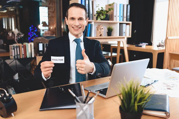 Realtor sitting at desk in office. Man is posing on camera with realtor sign. — Stockfoto